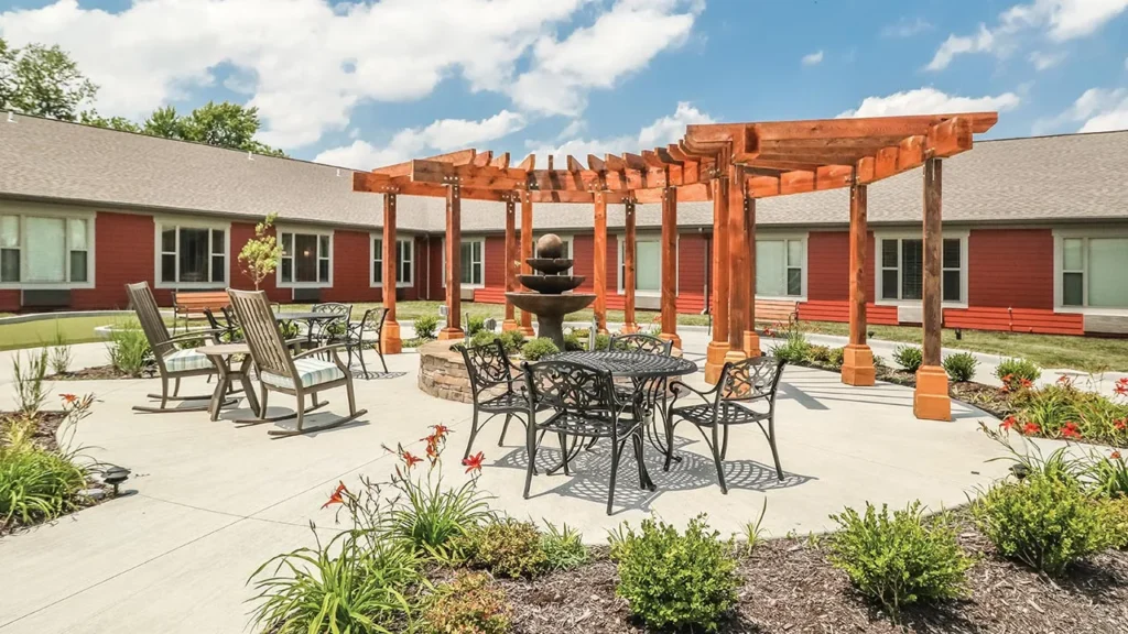 Outdoor courtyard with a wooden pergola over a circular stone fountain. Surrounding the pergola are metal tables and chairs on a concrete patio. Red, single-story building with several windows forms an L-shape around the courtyard. Landscaping includes flowers and bushes.