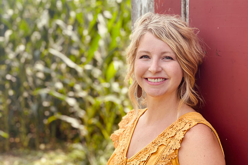 A woman with wavy blonde hair and a bright smile stands next to a red wall, wearing a yellow lace top. Behind her, tall green plants are visible, suggesting a rural or garden setting.