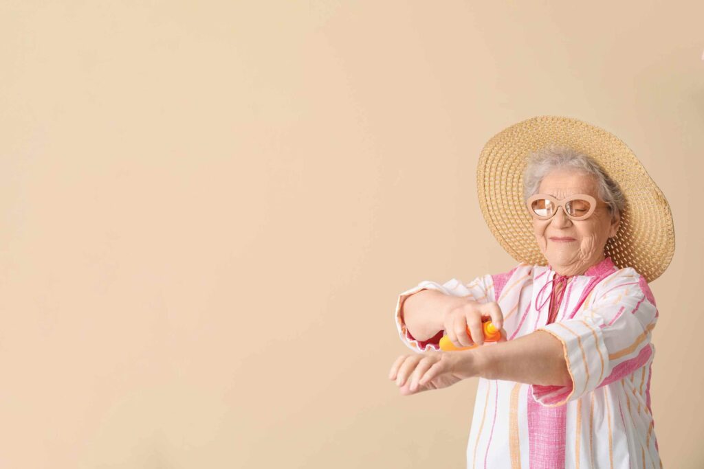 An elderly woman wearing a wide-brimmed straw hat and sunglasses is applying sunscreen to her arm. She is dressed in a light-colored, striped outfit and is set against a beige background.