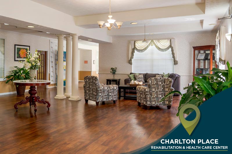 A spacious lobby area with wooden floors, floral arrangements, and cozy armchairs around a small table. Large windows with curtains allow natural light in. A sign reads "Charlton Place Rehabilitation & Health Care Center.