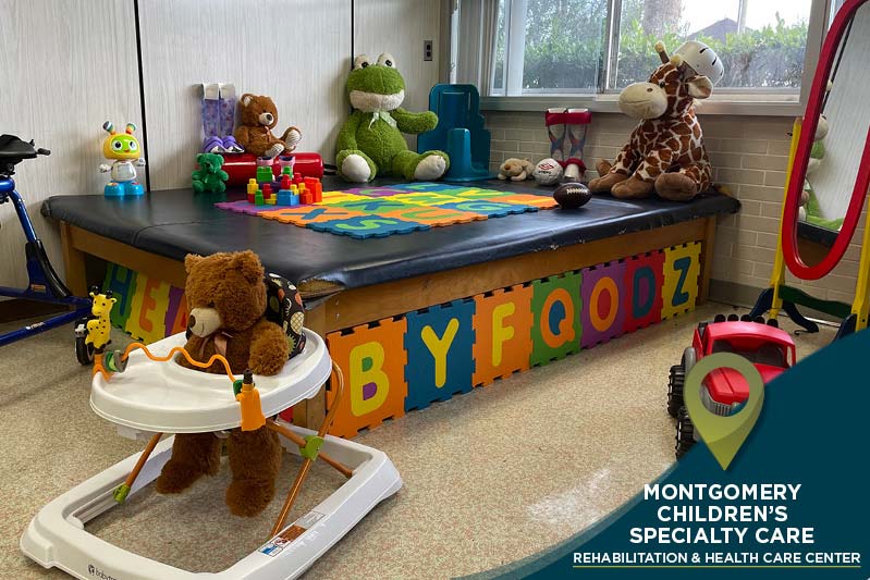 A children's playroom with a padded area surrounded by foam alphabet mats. Stuffed animals, including a teddy bear, giraffe, and frog, are placed around. Toys are scattered nearby. A sign reads "Montgomery Children's Specialty Care Rehabilitation & Health Care Center.