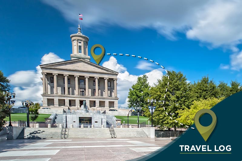 A historic government building with grand columns and a central dome, under a partly cloudy sky. A location marker hovers over the building, with a path leading to a second marker in the foreground. Text at the bottom right reads "TRAVEL LOG.