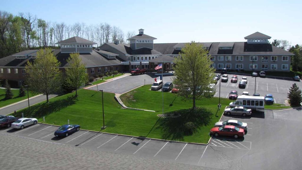A large building surrounded by greenery with a spacious parking lot in the foreground. Several vehicles, including cars and a bus, are parked. The building has multiple sections with peaked roofs, and a flagpole with the American flag stands near the entrance.