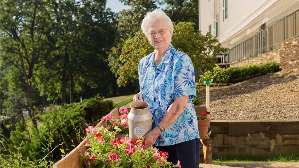 An elderly woman with short white hair, wearing a blue floral shirt and pants, smiles while watering vibrant pink and white flowers in a garden. She holds a silver watering can. The setting is outdoors with trees and a building in the background.