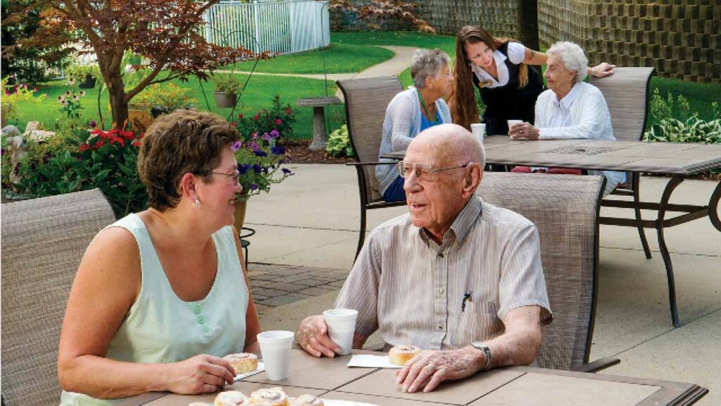 A group of elderly people enjoys a sunny day sitting at outdoor tables in a garden setting. A woman engages in conversation with an elderly man, both holding coffee cups, while other seniors chat in the background near well-maintained landscaping and trees.