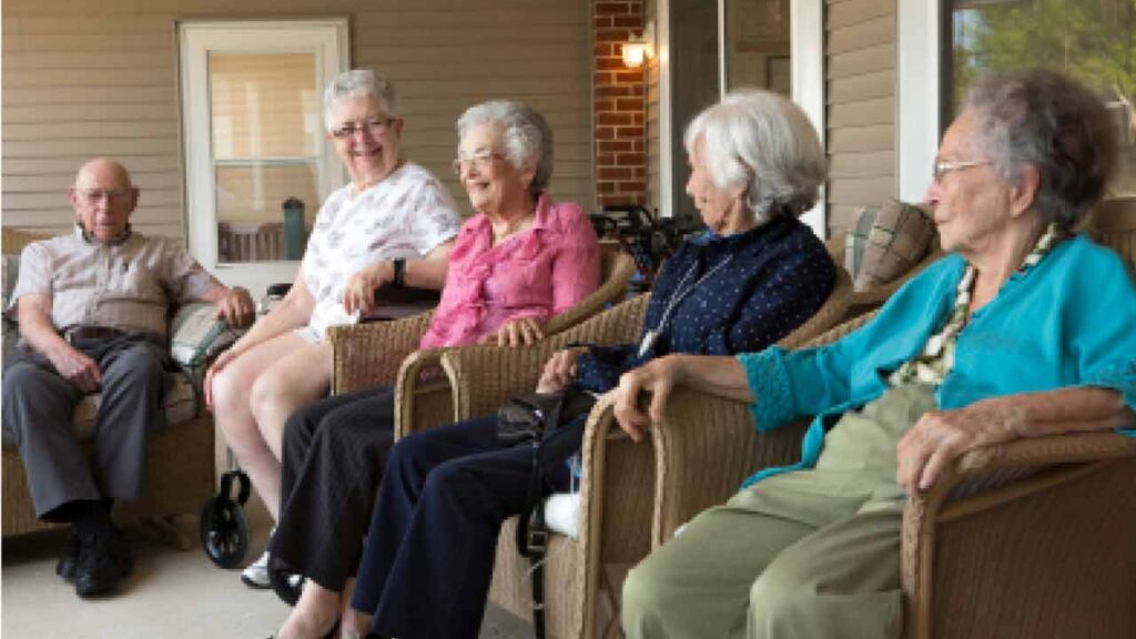 Five elderly individuals sit and smile on a porch, engaged in conversation. They are seated in wicker chairs, with a beige wall and a white door in the background. The setting appears relaxed and sociable.