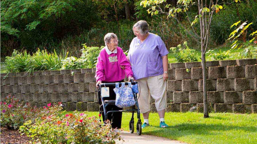 Two elderly women are walking along a garden path, one using a walker with a blue floral bag attached. One woman, in a pink jacket, is assisted by another woman in a purple shirt and capri pants. They are surrounded by greenery and flowers.