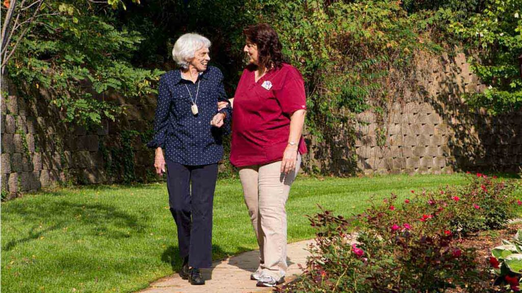 An elderly woman with white hair, wearing a blue shirt and dark pants, is walking arm-in-arm with a younger woman in a maroon shirt and beige pants. They are in a garden, smiling at each other, surrounded by greenery and blooming flowers.