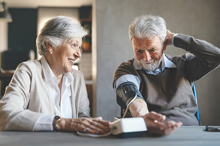 An elderly man uses a blood pressure monitor while sitting at a table. A smiling elderly woman sits next to him.