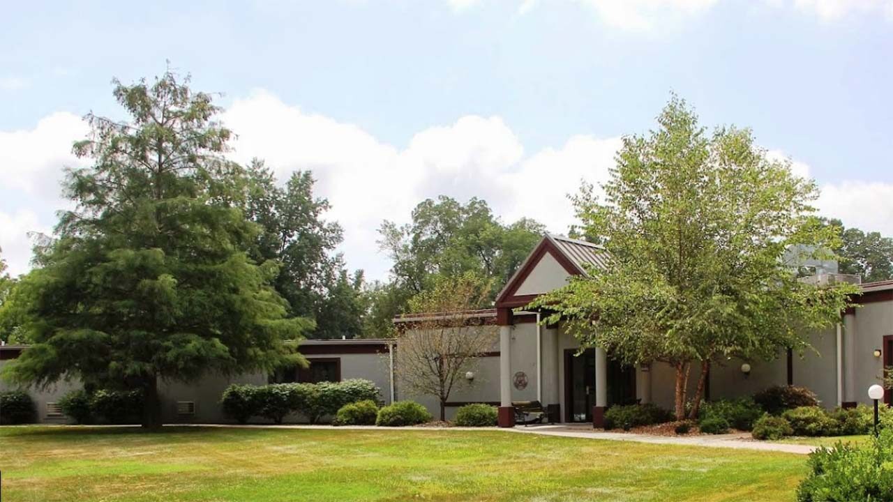 A one-story building surrounded by green trees and shrubs. The structure has a flat roof with a red triangular section above the entrance. A well-maintained grassy area is in the foreground, under a cloudy sky.