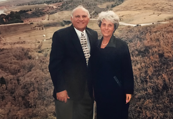 An elderly couple stands together, smiling, in front of a scenic landscape with hills and trees. The man is in a suit with a patterned tie, and the woman is wearing a dark coat. The background features fields and a winding road.