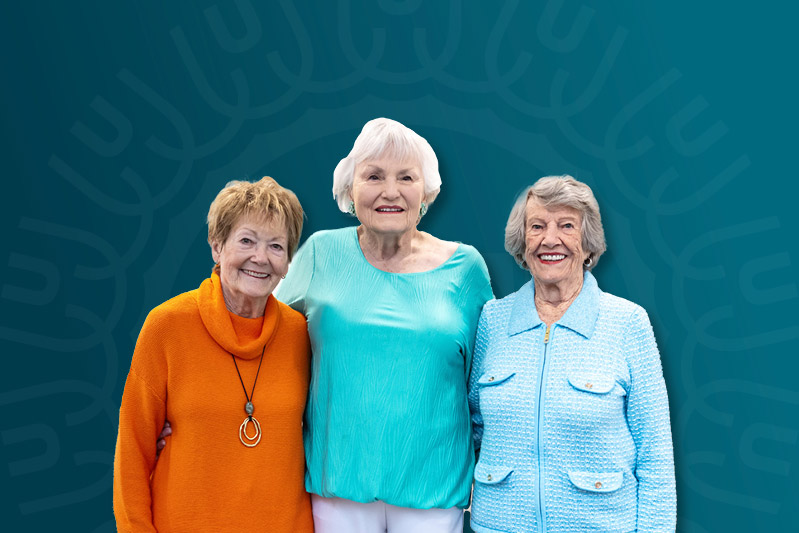 Three elderly women stand together, smiling, against a teal backdrop with a subtle pattern.