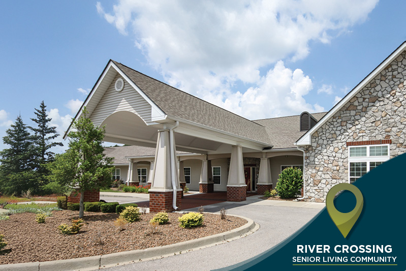 The image shows the entrance of the River Crossing Senior Living Community. It features a large covered driveway, brick accents, and landscaped gardens under a partly cloudy sky. A logo with a map marker and the community's name is in the bottom right corner.