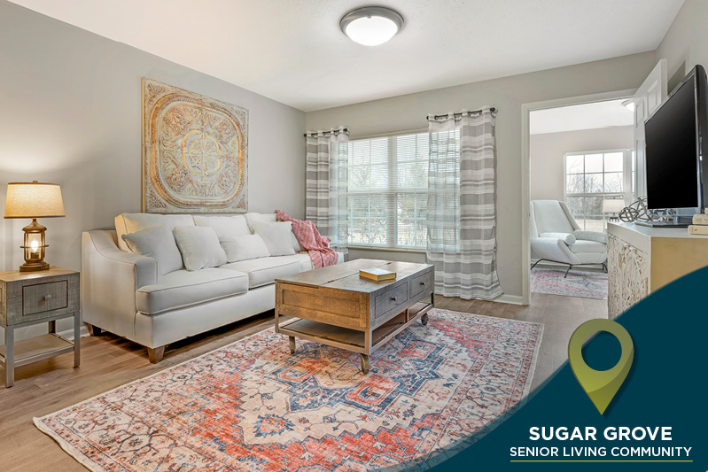 Bright living room with a white sofa, wooden coffee table, and patterned rug. A floor lamp stands beside the sofa, and a large window offers natural light. An open doorway leads to another room. Text reads: "Sugar Grove Senior Living Community.