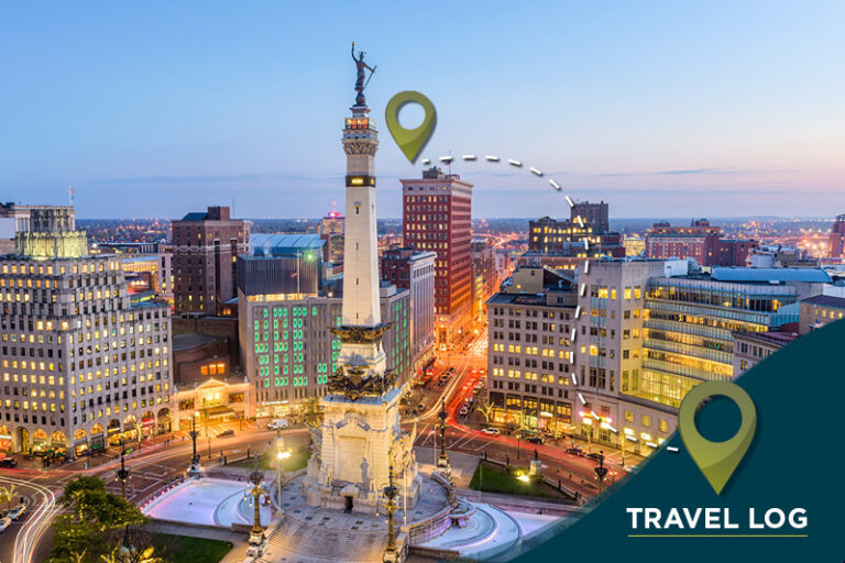 Aerial view of a city's monument circle at twilight, featuring a tall monument surrounded by illuminated buildings. The image includes map pin graphics and text reading "Travel Log" in the bottom corner.