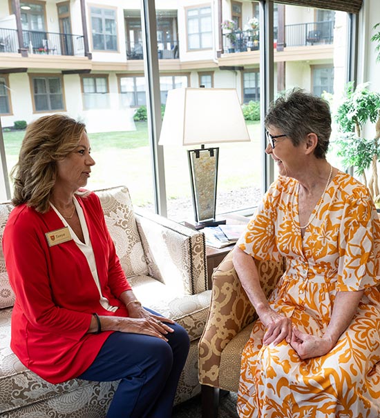 Two women are sitting in a well-lit room with large windows. One woman, in a red top, is speaking to another woman in a yellow patterned dress. They are seated on a sofa and chair, respectively, engaging in conversation.