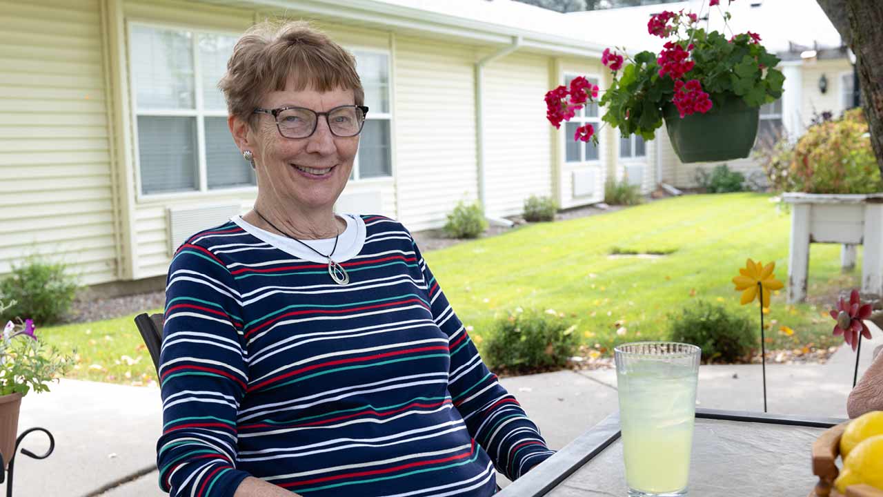 A woman with glasses, wearing a striped shirt, sits at an outdoor table with a glass of lemonade. She smiles, surrounded by flowers and greenery. A building with windows is in the background.