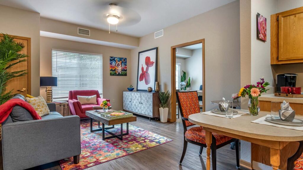 A cozy living room with a gray sofa, red armchair, and colorful rug. The dining area has a small wooden table with chairs. Various plants and vibrant art decorate the walls. A ceiling fan is on, and sunlight streams through a window with blinds.