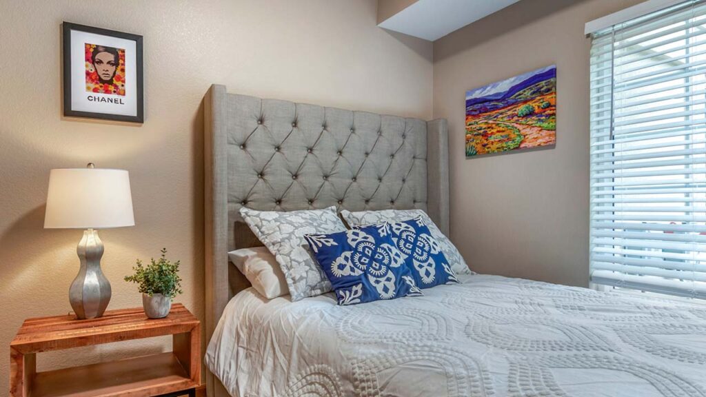 A cozy bedroom with a tufted gray headboard and a white quilted bedspread. Decorative blue and white pillows are on the bed. A wooden side table with a lamp and a plant is beside the bed. Colorful artwork and a Chanel print are on the walls.