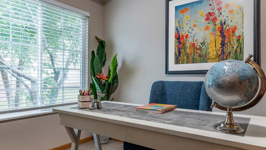 A well-lit office features a desk with a gray mat, globe, books, and a pen holder with colorful pens. A gray chair sits behind the desk. A vibrant floral painting hangs on the wall, and a large plant is positioned near a window with blinds.