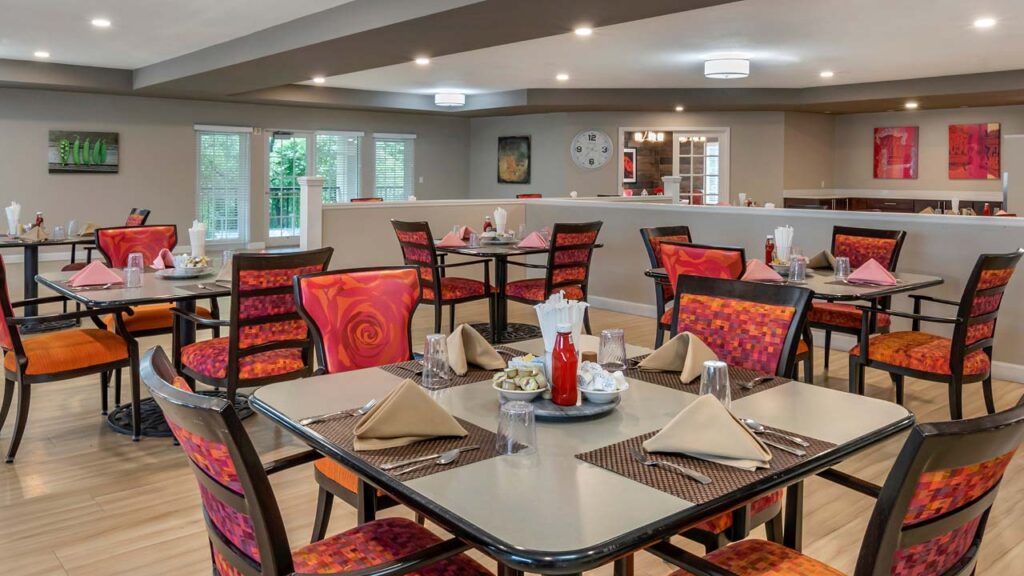 A modern dining area with square tables set for four. Each table has red and orange cushioned chairs, napkins, glasses, and condiments. The room features beige walls, abstract art, and large windows letting in natural light.