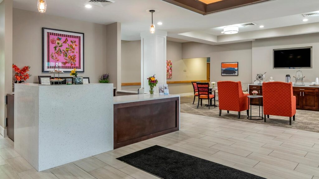 A modern lobby with a reception desk on the left, orange chairs around a small table on the right, and vibrant artwork on the walls. The room features neutral tones with splashes of color from decor and lighting fixtures.