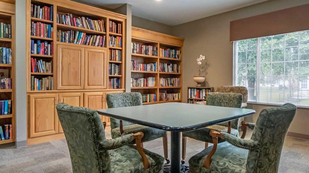 A cozy library room with a square table and four green upholstered chairs. Wooden bookshelves filled with colorful books line the walls. A large window allows natural light to enter, and a potted plant decorates a corner shelf.