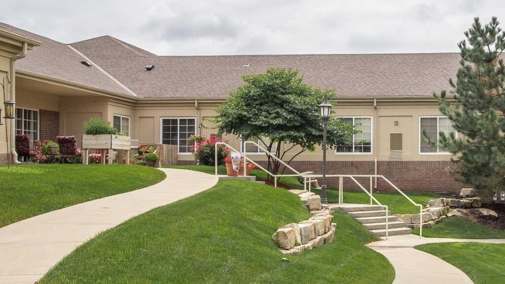 A landscaped garden with a winding path leads to a building. The path is lined with flowers, shrubs, and small stone walls. A mature tree stands near the stairs with a handrail. The building is beige with large windows and a grey roof.