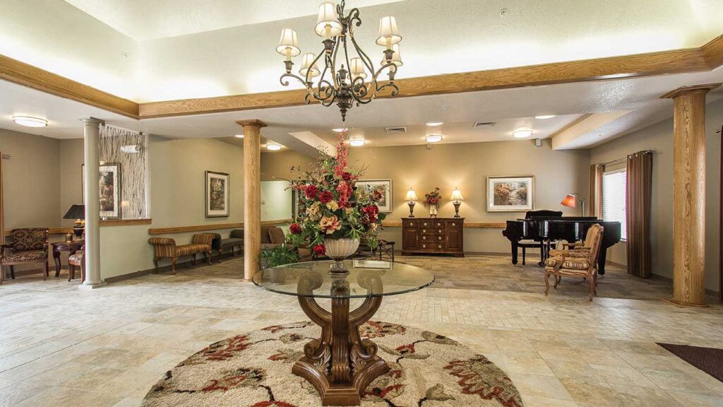 Spacious room with a round glass table featuring a large floral arrangement in the center. There are ornate chairs, a black piano, and framed artwork. The room is well-lit with chandeliers and has a beige and cream color scheme.