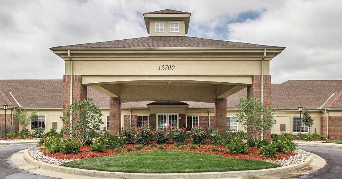 Large building with a central entrance featuring a covered driveway. The front has decorative shrubs, flowers, and a circular drive. The building number 