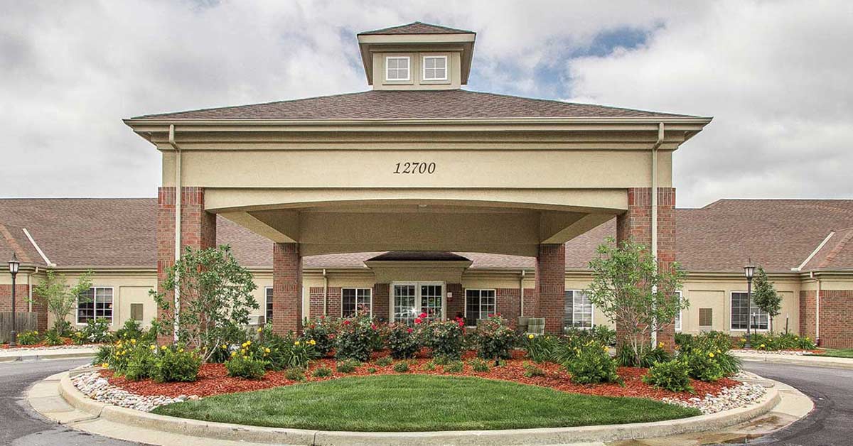 Large building with a central entrance featuring a covered driveway. The front has decorative shrubs, flowers, and a circular drive. The building number "12700" is displayed above the entrance. The sky is partly cloudy.