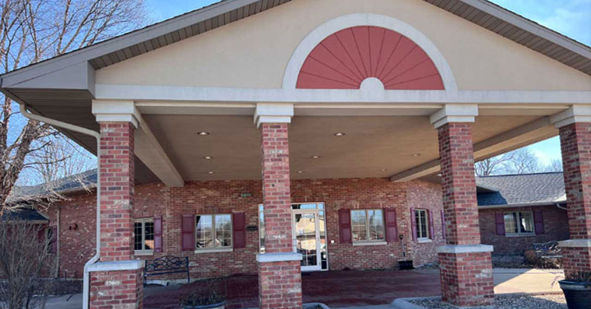 A brick building with a covered entrance supported by four columns. The entrance features a semicircular decorative element above the door. Bushes and a bench are visible on the left side, and the sky is clear.