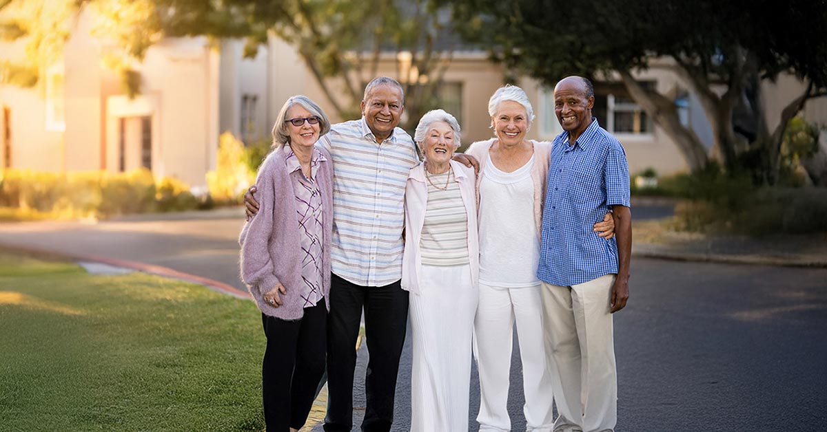 A group of five older adults, three women and two men, stand closely together outside on a sunny day. They are smiling, with trees and buildings in the background. They all wear casual, light-colored clothing.