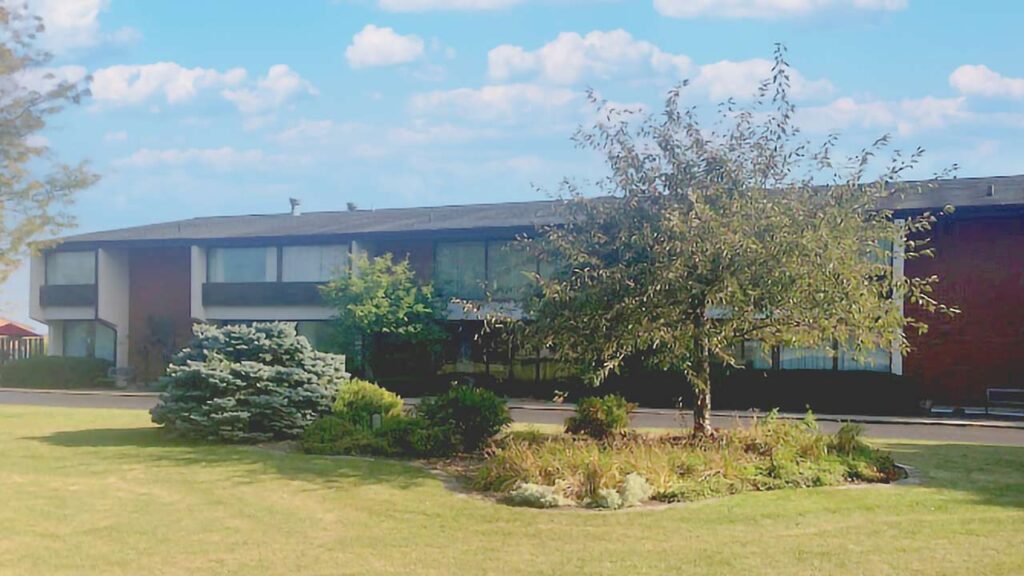 A two-story brick building with large windows stands behind a grassy area. In the foreground, there is a landscaped garden with a tree and various shrubs. The sky above is blue with scattered clouds.