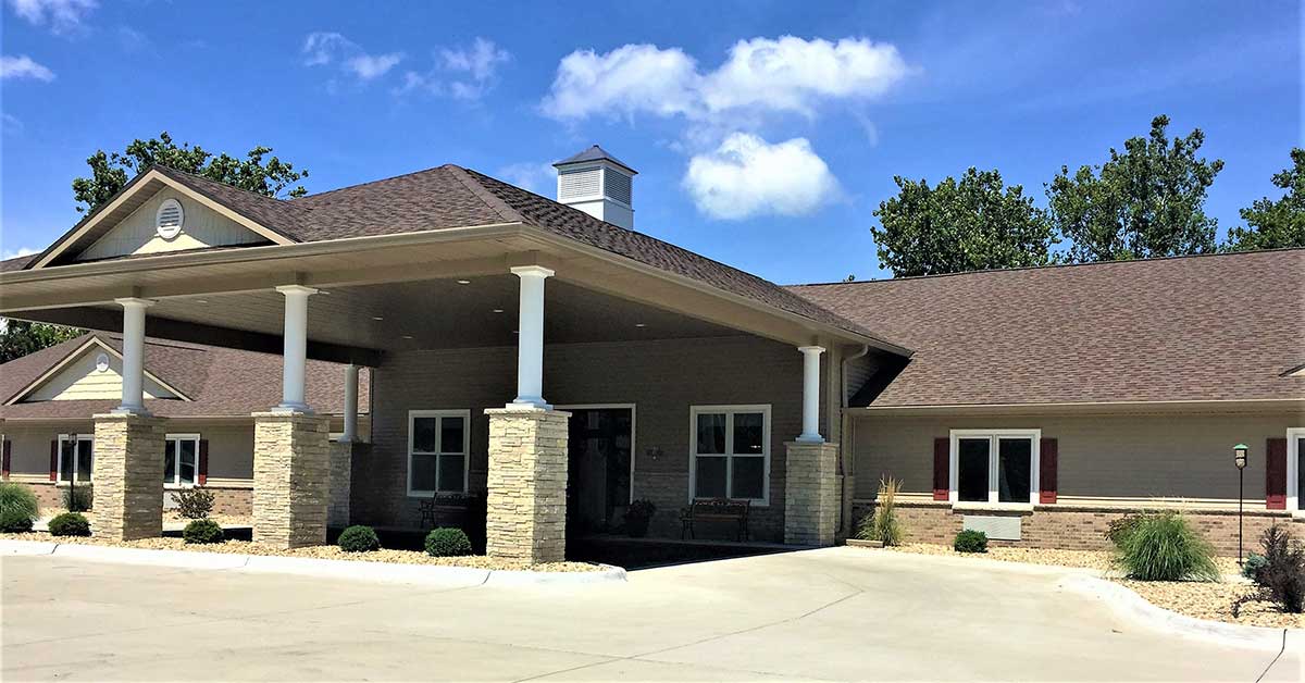 A single-story building with a covered entrance supported by columns. The exterior features beige siding, stone accents, and white trim. There are small shrubs and plants around the entrance, and a clear blue sky above.