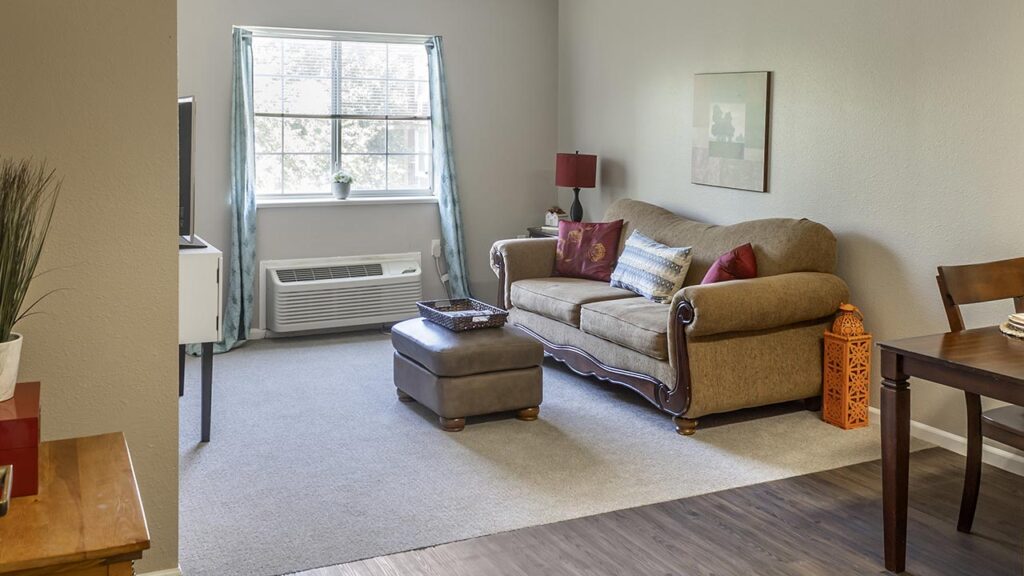Cozy living room with a brown couch and decorative pillows, an ottoman, a wall-mounted A/C under a sunlit window with blue curtains, and a small table with a red lamp. The room has neutral walls and a mix of carpet and wood flooring.