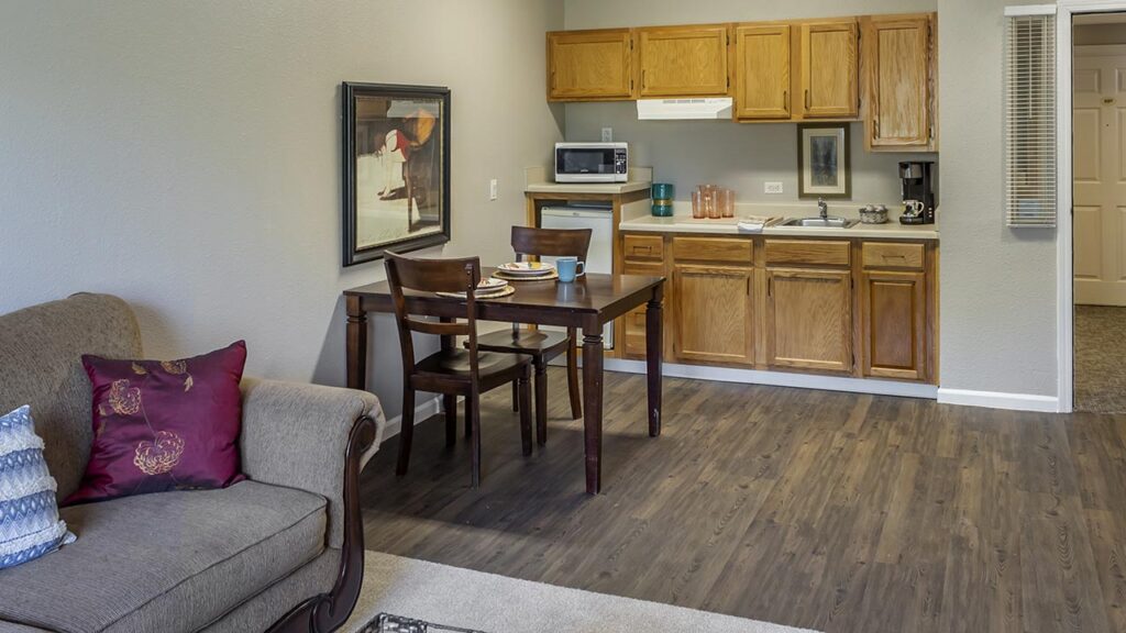 A cozy living space features a gray sofa with decorative pillows, a dark wooden dining table with two chairs, and a compact kitchen with wooden cabinets, a microwave, and a coffee maker. The floor is covered with light wood laminate.