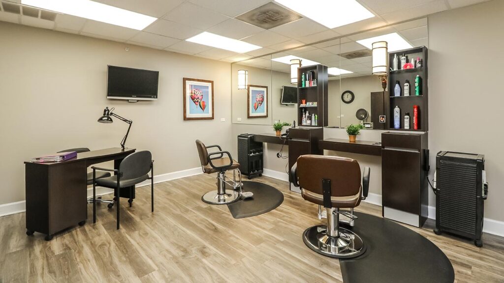 A modern hair salon interior featuring two styling chairs facing a mirrored wall with shelves of hair products. A small desk with a chair and lamp is on the left. Light wood flooring and framed art enhance the neutral-toned space.