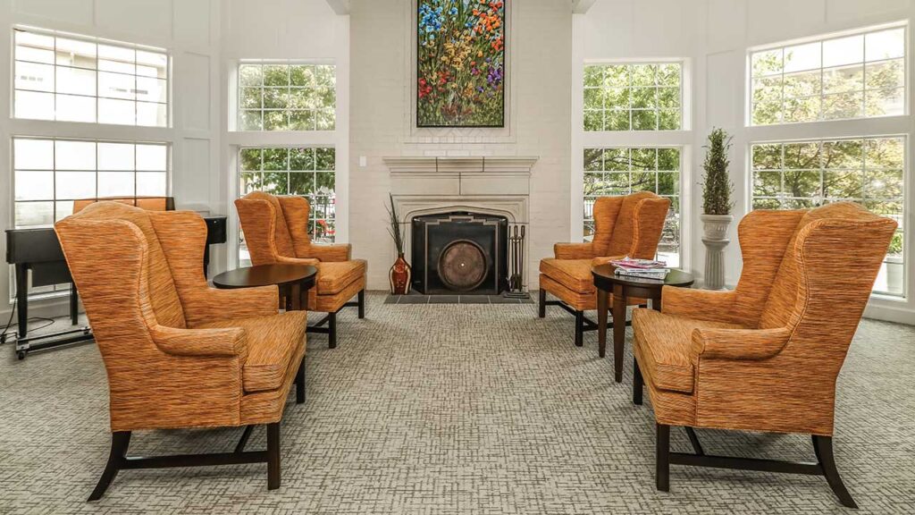 A cozy lounge area with four orange armchairs surrounding a circular wooden coffee table. A fireplace with a colorful painting above it is centered on the back wall. Large windows let in natural light.