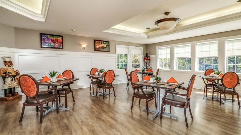 A bright dining room with wooden flooring, featuring six tables set with orange napkins. Each table has two chairs. Large windows allow natural light in, and paintings decorate the walls. A decorative chef statue stands in the corner.