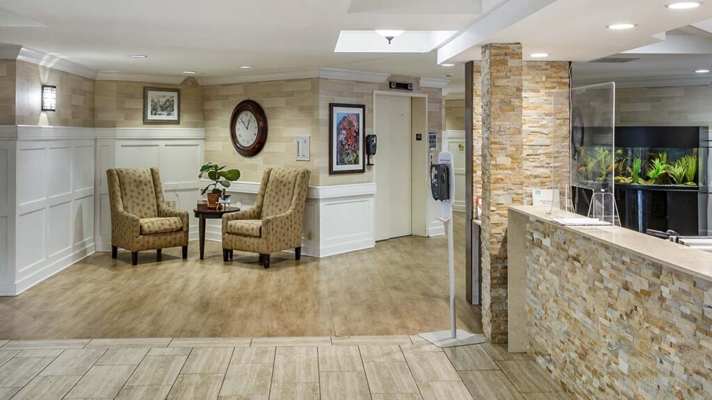 A welcoming lobby area with two patterned armchairs around a small table, beneath a clock and artwork. The room features wood-paneled walls and flooring, a reception desk with stone detailing, and an aquarium in the background.