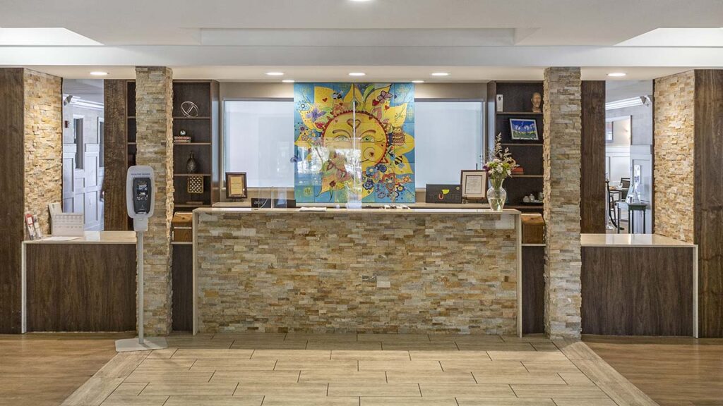 A hotel reception desk features a stone facade and is flanked by two stone columns. Behind the desk is a vibrant sun-themed artwork. The area is well-lit and has a hand sanitizer dispenser on the left.