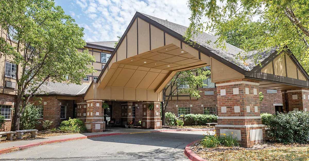 A brick building with Tudor-style architectural features has a covered entrance supported by brick columns. Trees and shrubs surround the entrance, and a stone bench is visible on the left. The sky is partly cloudy.