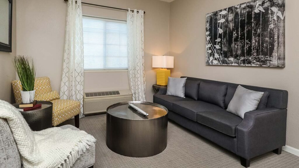 A cozy living room with a gray couch, patterned chairs, and a large round coffee table. The window is adorned with sheer curtains. A black and white abstract painting hangs on the wall, and a lamp with a yellow base sits nearby.