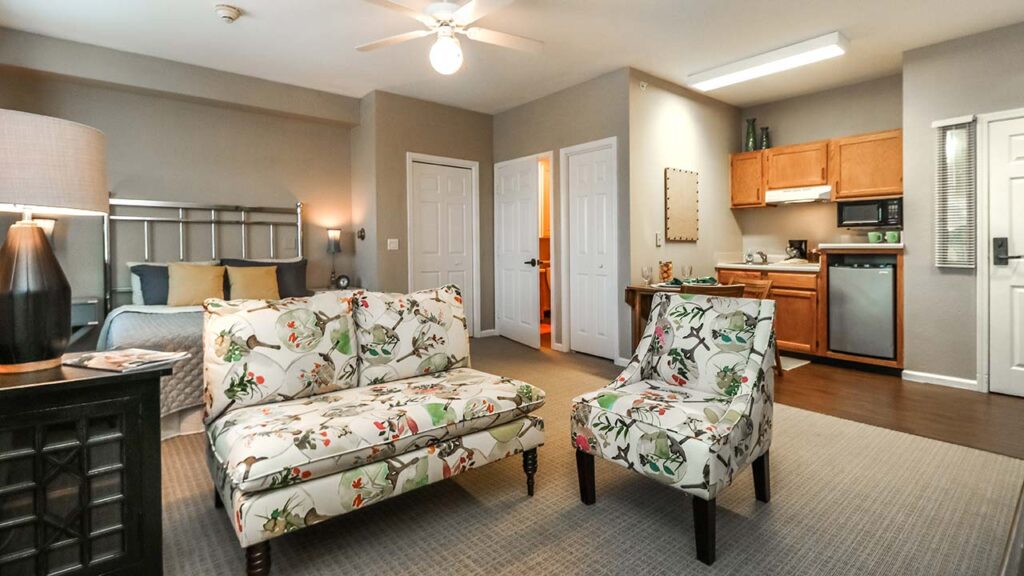 A cozy studio apartment featuring floral-patterned armchairs and a loveseat facing a dark wood side table. A bed is in one corner, and a small kitchen area with wooden cabinets and stainless steel appliances is in another. A ceiling fan is above.