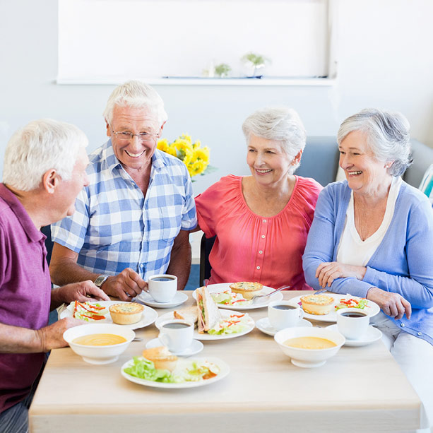 Four elderly people sit around a table, enjoying a meal together. Plates with salads, sandwiches, soup, and cups of coffee are arranged on the table. They smile and converse in a bright room with flowers in the background.