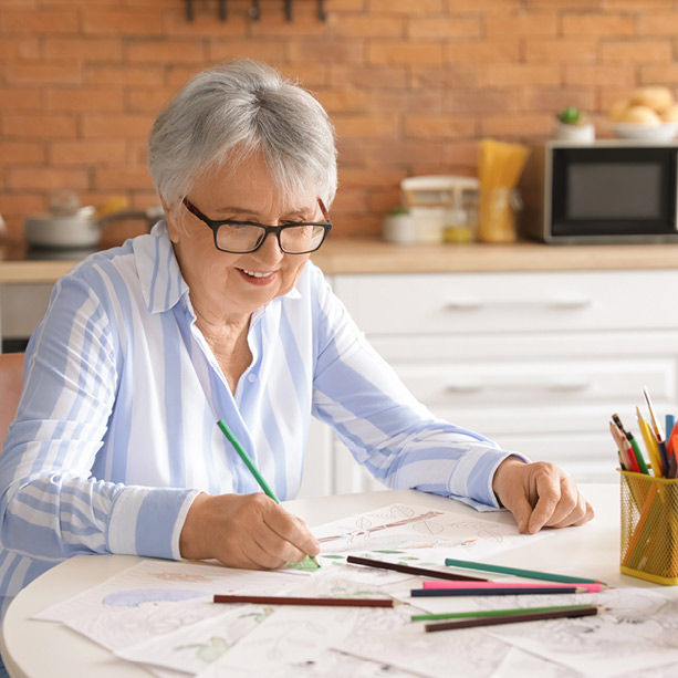 Elderly woman with glasses and short gray hair is sitting at a table drawing with colored pencils. She's wearing a striped shirt and smiling slightly, with a kitchen background featuring a brick wall and shelves.