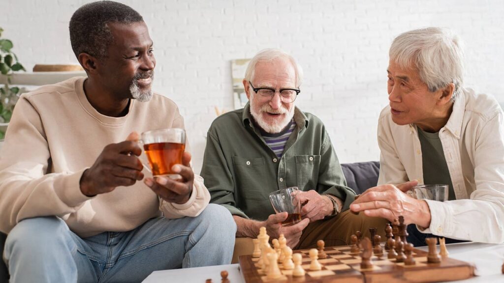 Three elderly men sitting together around a chessboard, engaged in conversation. One holds a cup of tea, another a glass, and the third gestures while speaking. The setting is cozy, with a light-colored brick wall in the background.