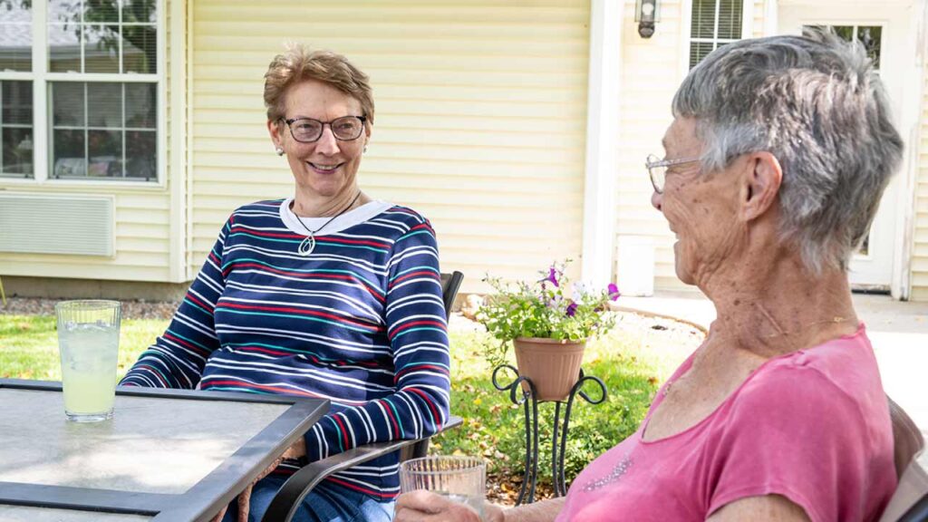 Two older women sit outside at a table with drinks. One woman wears a striped shirt and the other a pink shirt. They appear to be talking and smiling, with a cream-colored building and potted plants in the background.