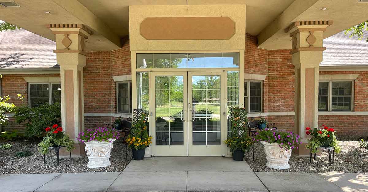 Entrance of a building with glass double doors and large windows. Brick walls flank the entry, which is adorned with potted plants and flowers on either side. The roof is supported by decorative pillars. A path leads up to the entrance.